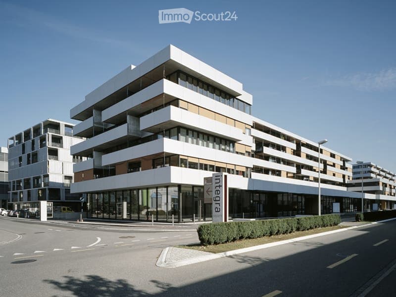 Modern multi-story building with white and brown facade, large windows, and a sign reading "Lageplan" in front.