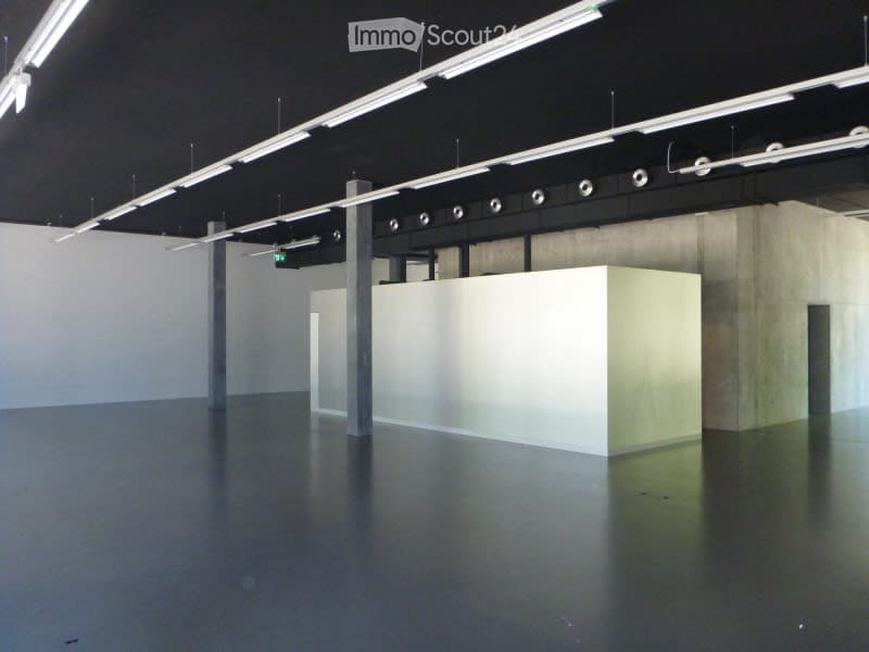 Empty, unfurnished room with a polished concrete floor, white walls, and a single white partition. Exposed metal beams and industrial lighting fixtures are visible on the ceiling.