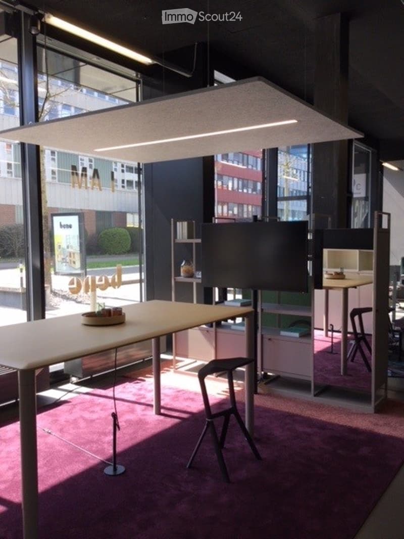 Modern office space with large windows, a long wooden table, a stool, a screen, shelves, and chairs.