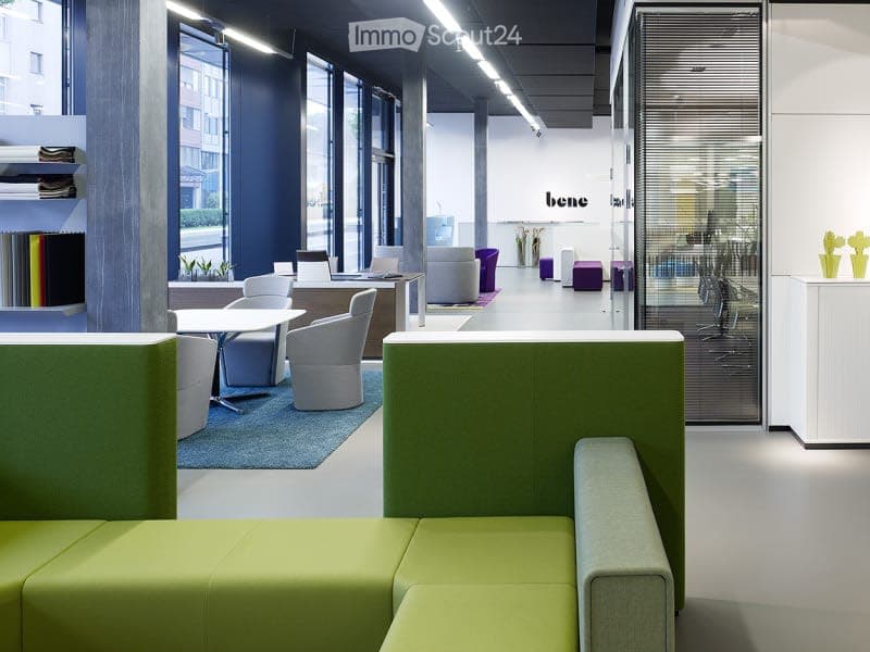 Modern office space with large windows, green and grey furniture, white walls, and a mix of seating and work areas.
