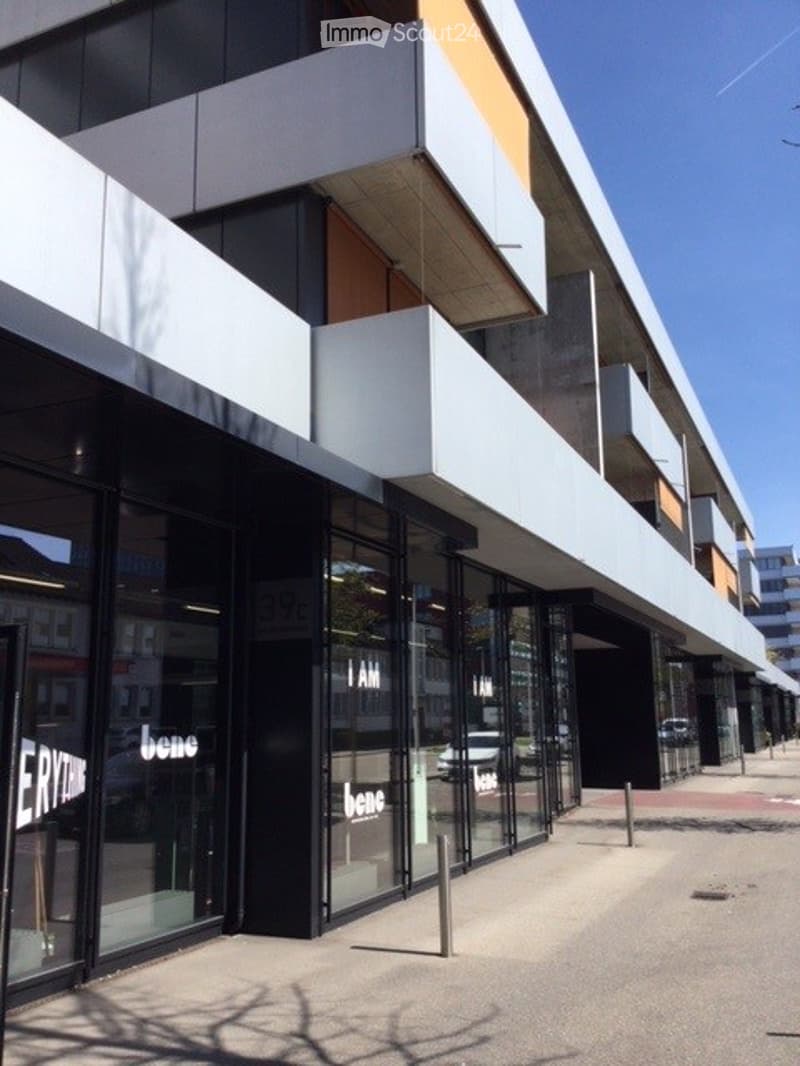 Modern commercial building with large glass storefronts and white balconies, featuring the word "bene" on the windows.