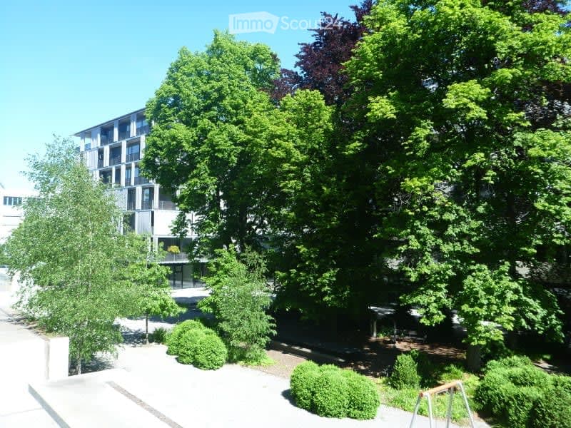 A modern residential building surrounded by lush green trees and neatly trimmed bushes, with a paved walkway leading to the entrance.