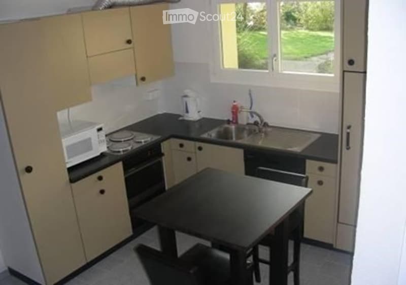 Compact kitchen with light wood cabinets, black countertop, microwave, oven, sink, and a small dining table with chairs.