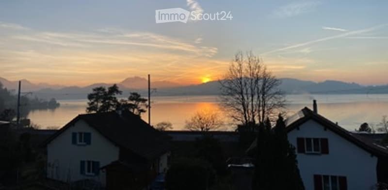 View of a serene lakeside scene with houses, mountains, and a sunset over the water.