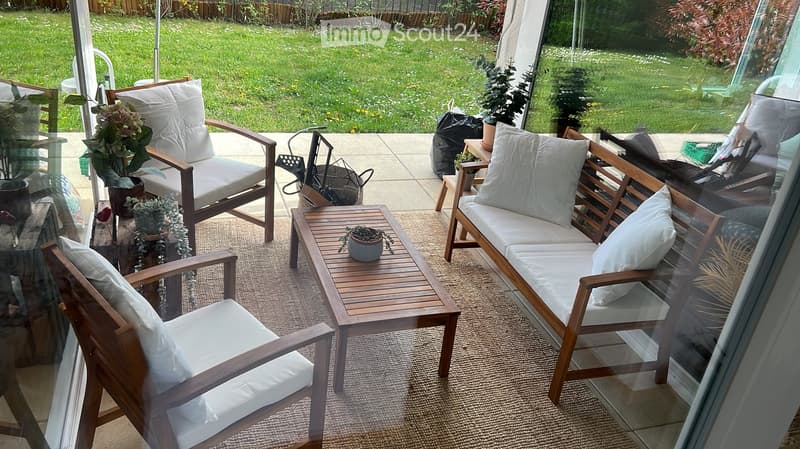A well-furnished balcony or terrace with wooden chairs, a sofa, a small table, and potted plants, overlooking a green garden.