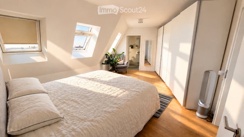 Bright, airy bedroom with wooden flooring, large windows, a bed with pillows, a chair, a plant, and a fan. The room has white walls and a modern aesthetic.