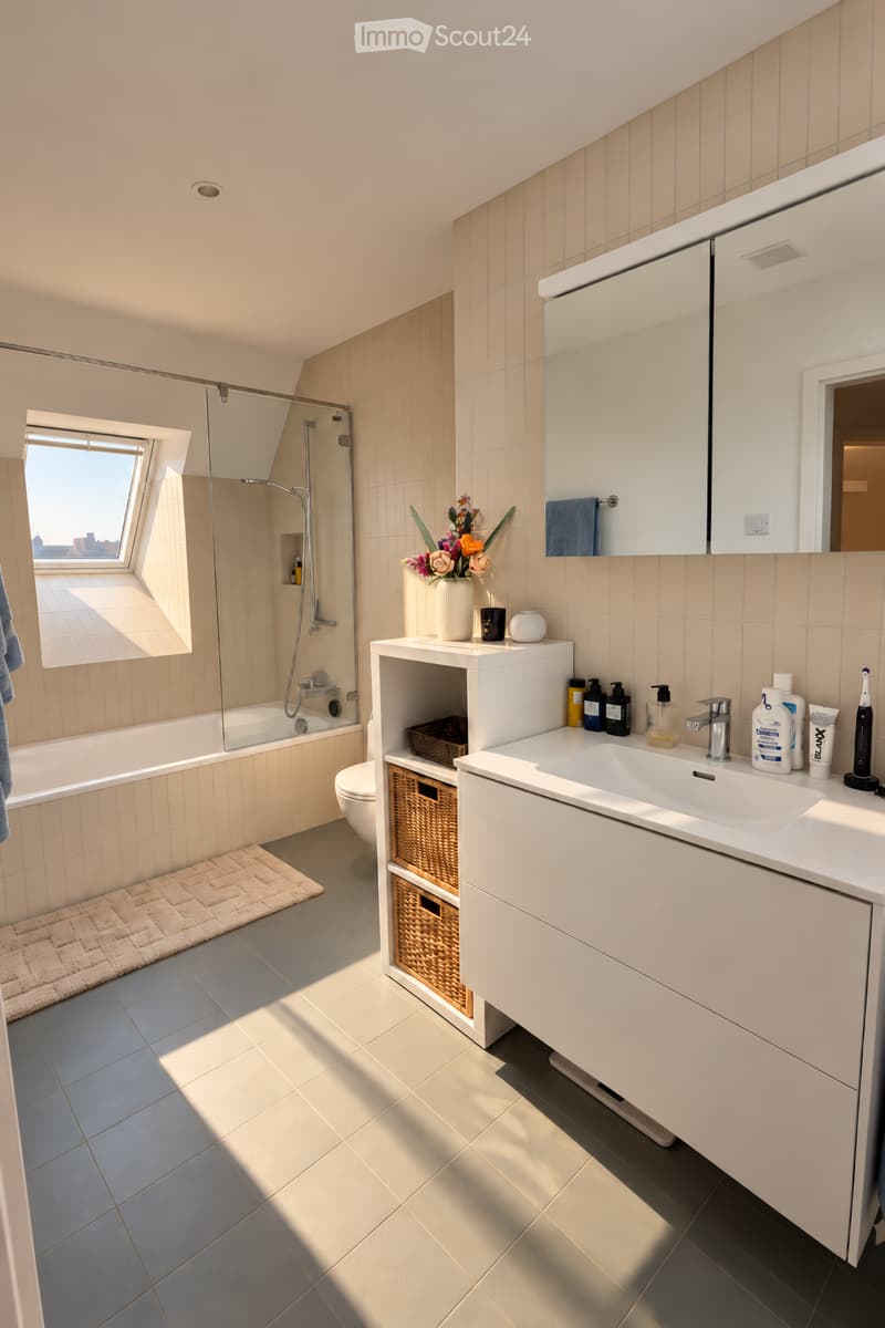Modern bathroom with a bathtub, sink, mirror, and toilet. Features a large window, wicker baskets, and a vase with flowers on the sink counter.