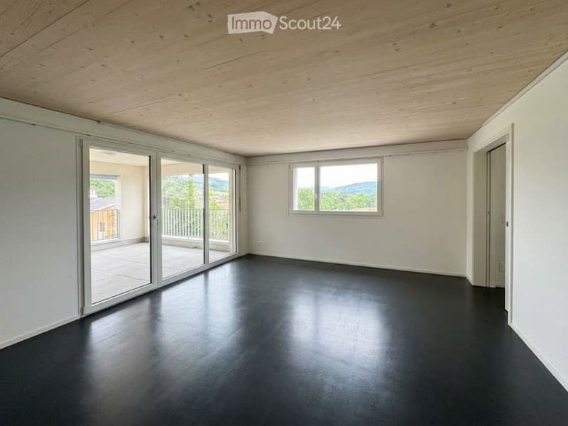 Empty room with white walls, dark flooring, large sliding glass doors leading to a balcony with a scenic view, and a window with a view of greenery and mountains.