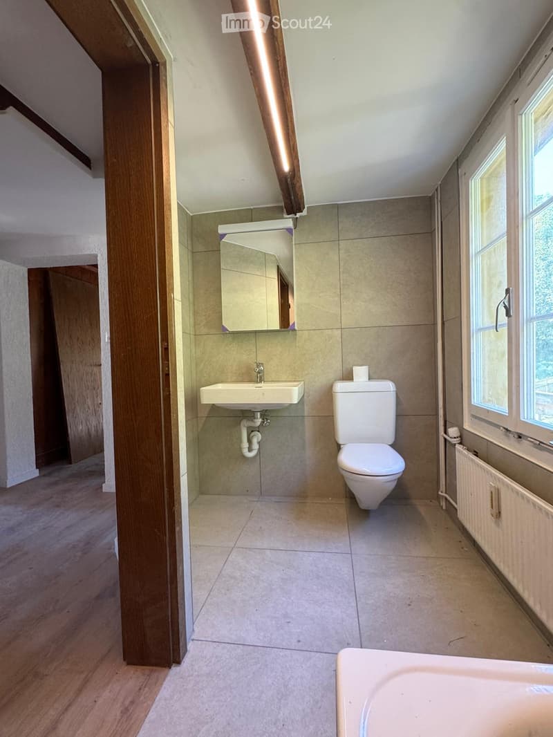Modern bathroom with beige tiles, a toilet, a sink, and a mirror. The room features wooden flooring and a window providing natural light.