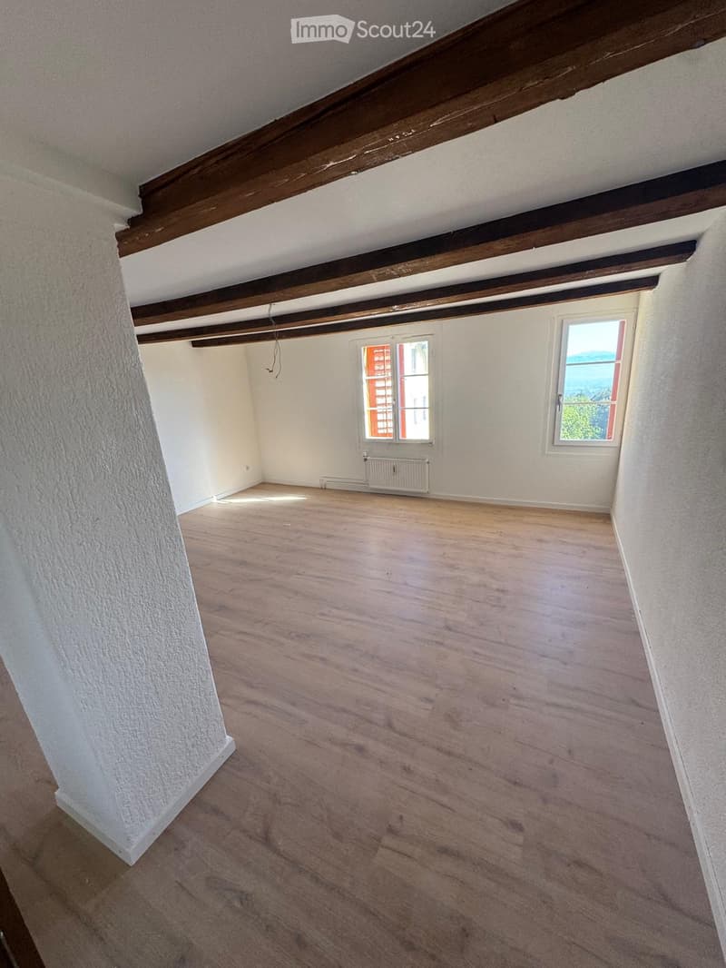 Empty room with wooden flooring, white walls, exposed wooden beams on the ceiling, and two windows offering natural light.