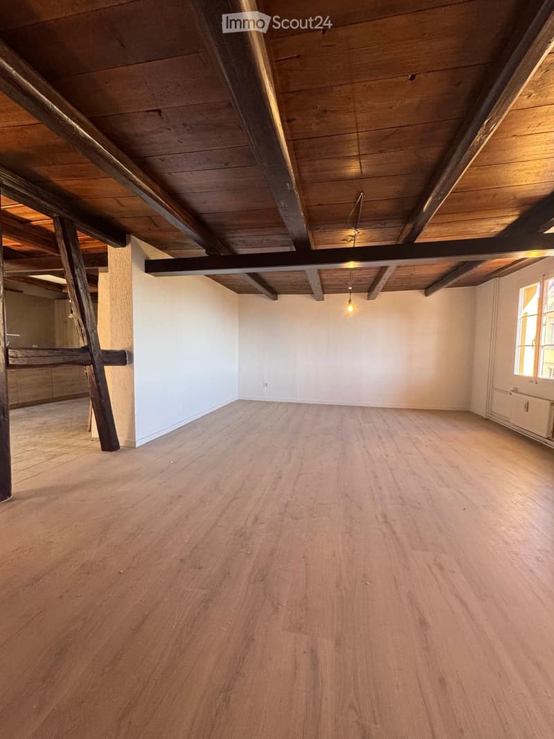 Empty room with wooden flooring, exposed wooden beams on the ceiling, white walls, and a single window letting in natural light.