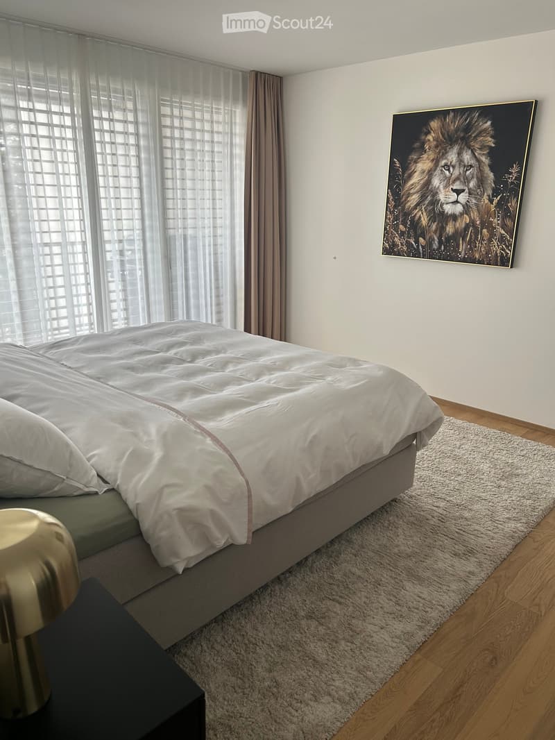 Modern bedroom with a large bed, nightstand, lamp, and a large painting of a lion on the wall. The room features wooden flooring and a white shaggy rug.