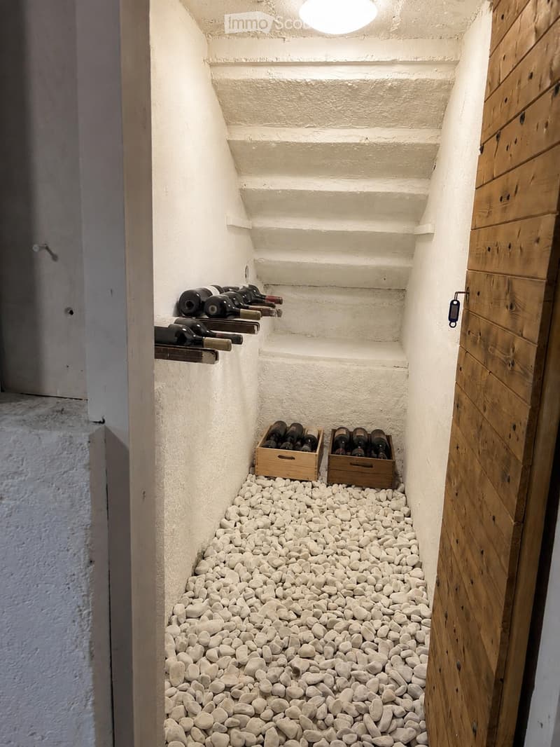 Narrow hallway with white textured walls, wooden door, and white pebble flooring. Wine bottles are stored on shelves and in wooden crates along the walls.