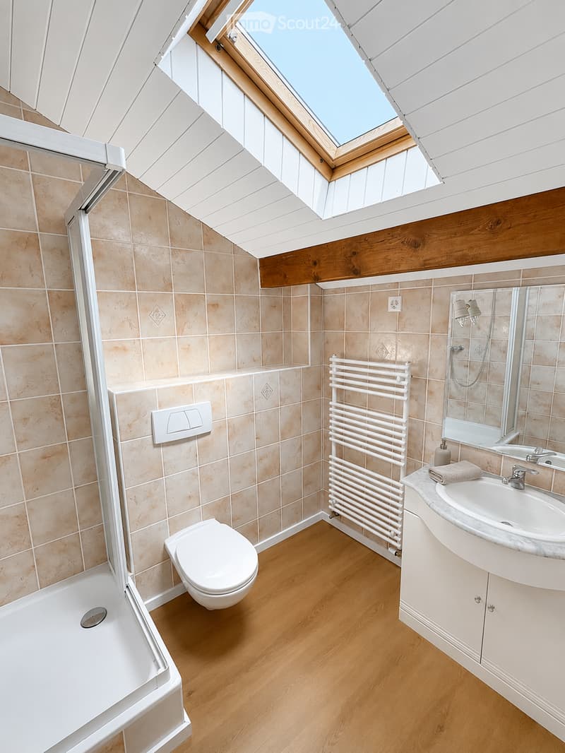 Modern bathroom with wooden flooring, tiled walls, a toilet, a sink with a mirror, a bathtub, and a white radiator. Features a skylight window and a towel on the sink counter.