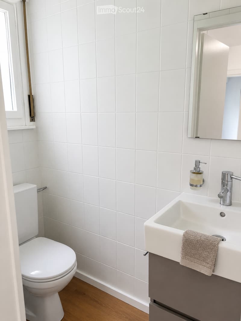 Modern bathroom with white tiled walls, a white toilet, a white sink with a modern faucet, a mirror above the sink, and a towel on the sink counter.