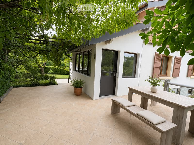 A tiled patio area with a wooden table, two benches, and a small potted plant, adjacent to a modern house with large windows and shutters.