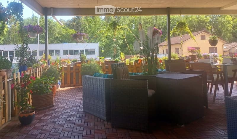 A covered outdoor patio with wooden furniture, potted plants, and a view of a garden and neighboring house.