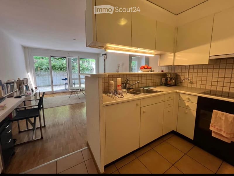 The image shows a modern kitchen with white cabinets, a sink, and a small dining area with a table and chairs. The kitchen features a tiled floor and a countertop with various appliances, including a coffee maker and a refrigerator. There is also a view o