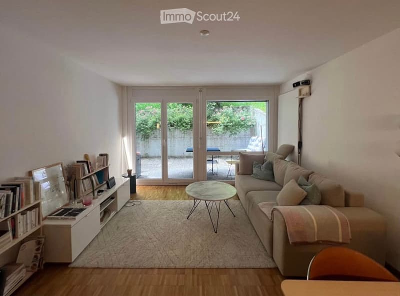 Bright living room with wooden flooring, large sliding glass doors leading to a balcony, a beige sofa with multiple cushions, a round coffee table, a bookshelf, and a small table with a chair.