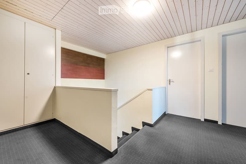 Empty hallway with carpeted floor, white walls, and a staircase leading upwards. Features a closed door on the right and a decorative panel on the left wall.