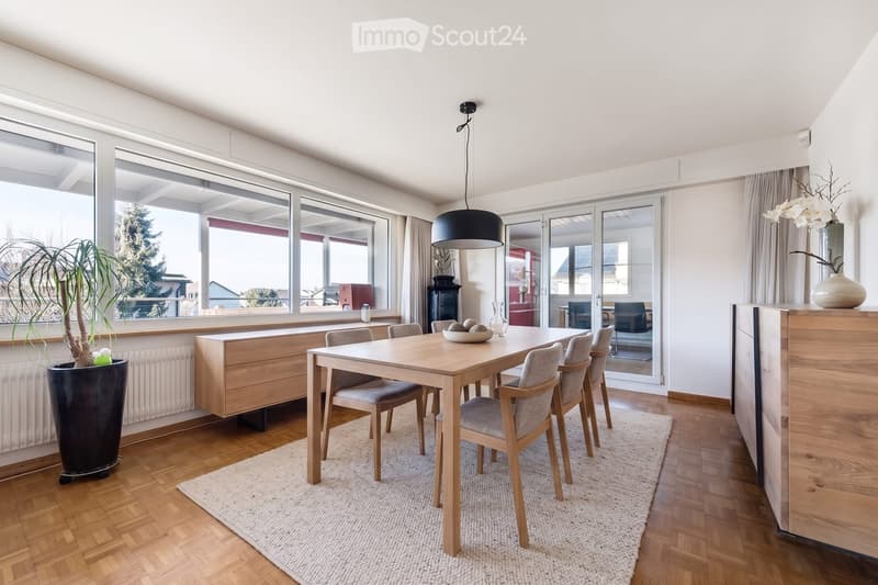 Bright dining area with a wooden dining table and six chairs, a pendant light, a large window with a view, and a potted plant near the window.