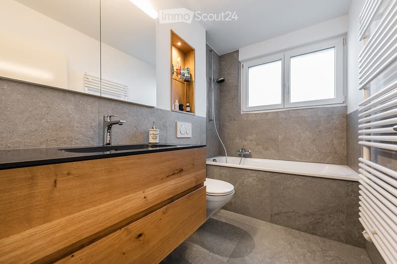 Modern bathroom with a wooden vanity, black sink, mirror, bathtub, toilet, and built-in shelving. Features include a radiator, a window, and a sleek faucet.