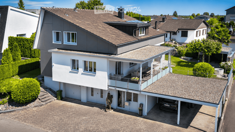 A modern two-story house with a garage, balcony, and well-maintained garden. The house features a combination of grey and white exterior walls, large windows, and a tiled roof.