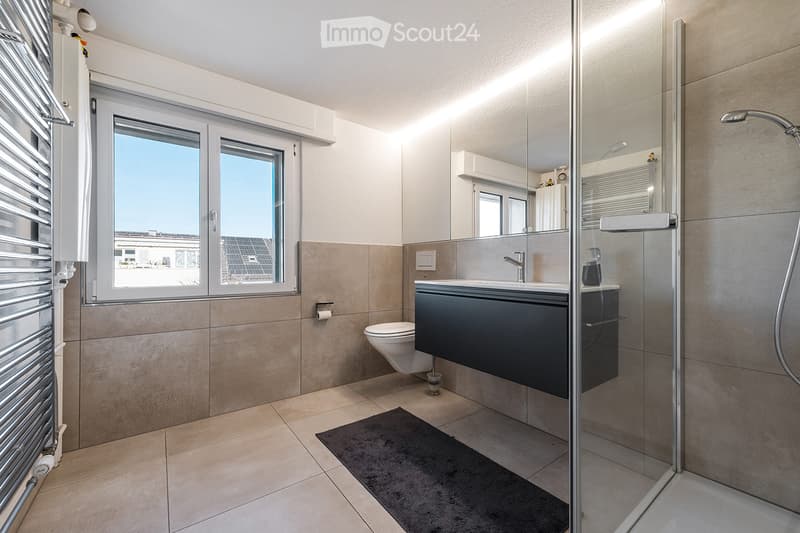Modern bathroom with a toilet, sink, and shower area. Features include a large mirror, a towel rack, and a radiator. The space is well-lit with natural light from a window.
