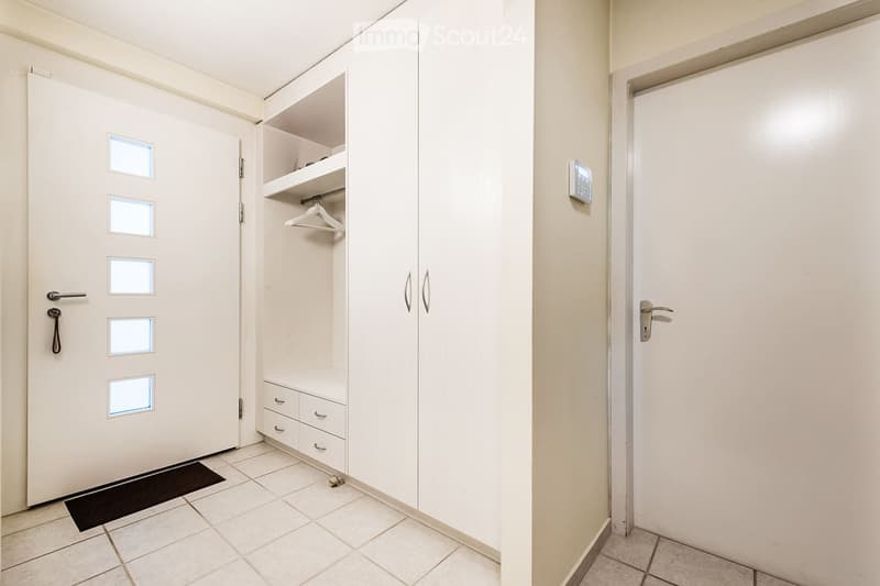 A clean, unfurnished hallway with tiled flooring, white walls, and a built-in closet with shelves and drawers. A door with frosted glass panels is visible on the left, and a small black doormat is placed in front of it. A digital thermostat is mounted on