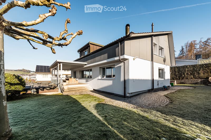 Modern two-story house with white and grey exterior, large windows, covered patio area, and well-maintained lawn.