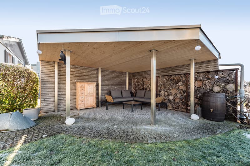 A modern outdoor patio with a wooden ceiling, stone walls, a grey sofa, coffee table, and a wooden cabinet.