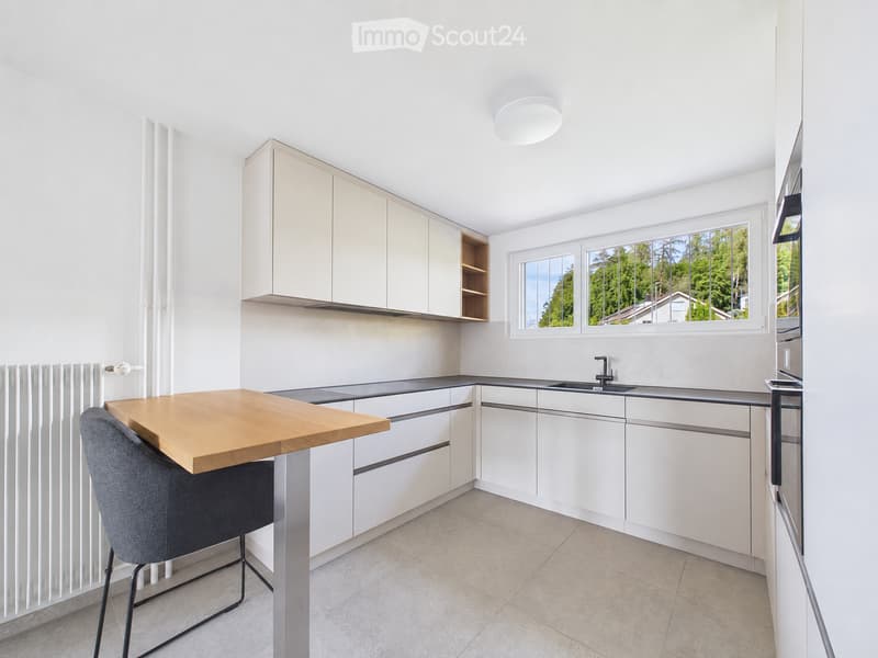 Modern kitchen with white cabinets, a wooden table, a grey chair, and a sink with a faucet. Large windows provide natural light and a view of greenery.