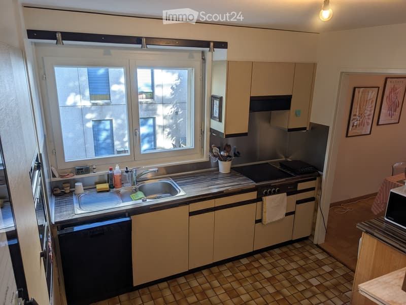 Modern kitchen with light-colored cabinets, stainless steel appliances, and a tiled floor. Features a sink, stove, microwave, and various kitchen items on the counter.