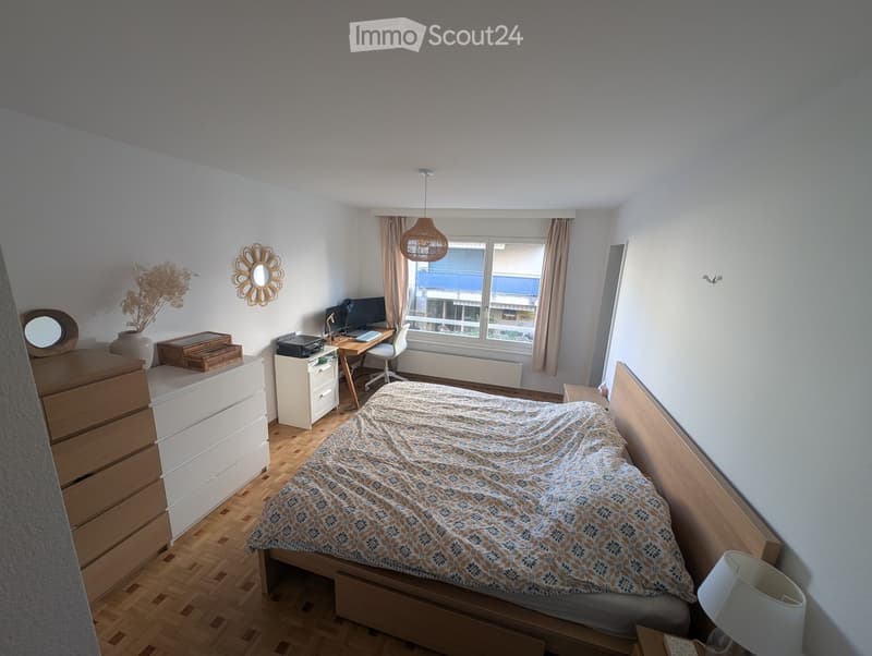 A well-lit bedroom with a wooden bed, nightstand, desk, and a window with curtains. The room features a patterned bedspread, a hanging pendant light, and decorative items on the dresser.