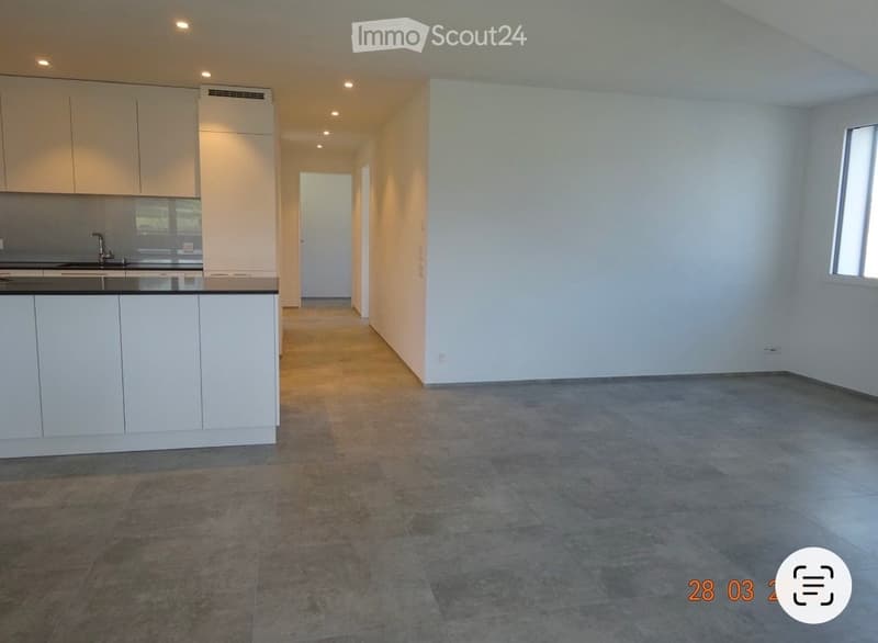 Empty room with tiled flooring, white walls, and a small kitchen area with cabinets and a sink visible in the background.