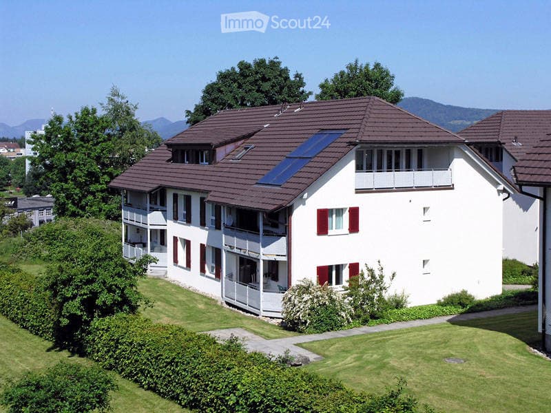A modern two-story residential building with white walls, red shutters, and a brown tiled roof. Features include multiple balconies, a solar panel on the roof, and a well-maintained lawn with hedges and trees in the foreground.