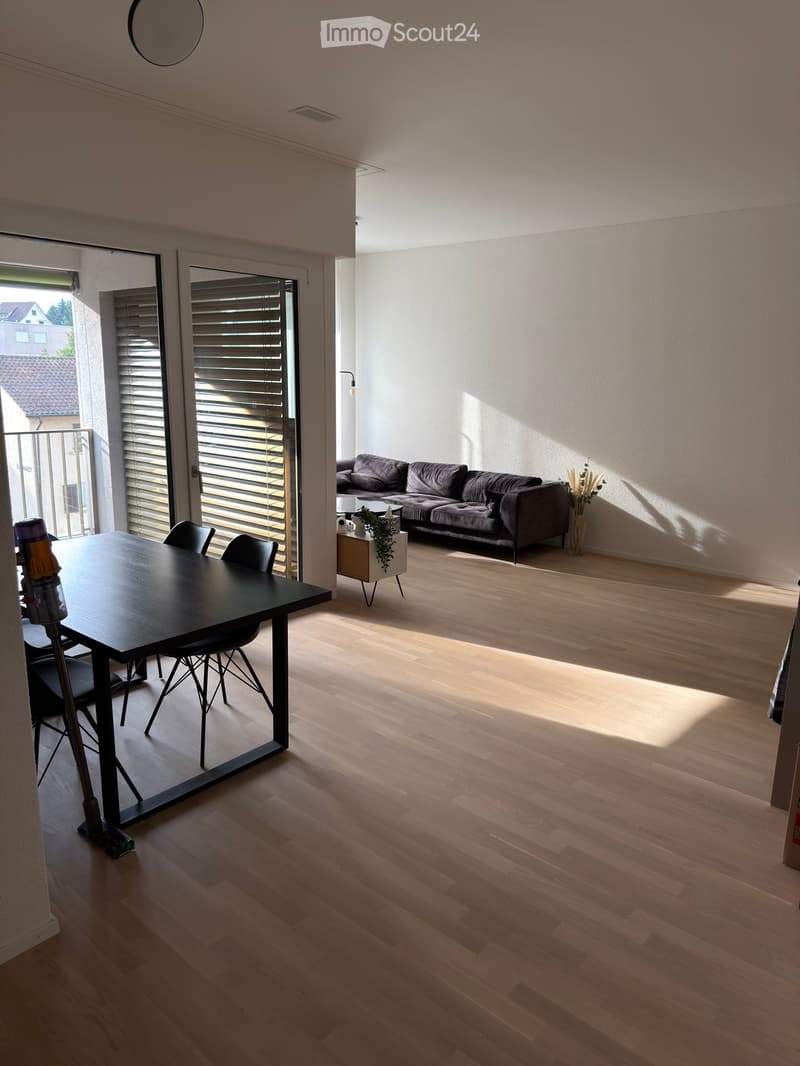Open-plan living and dining area with light wooden flooring, a grey sofa, a black dining table with chairs, and a small side table with a plant and vase.