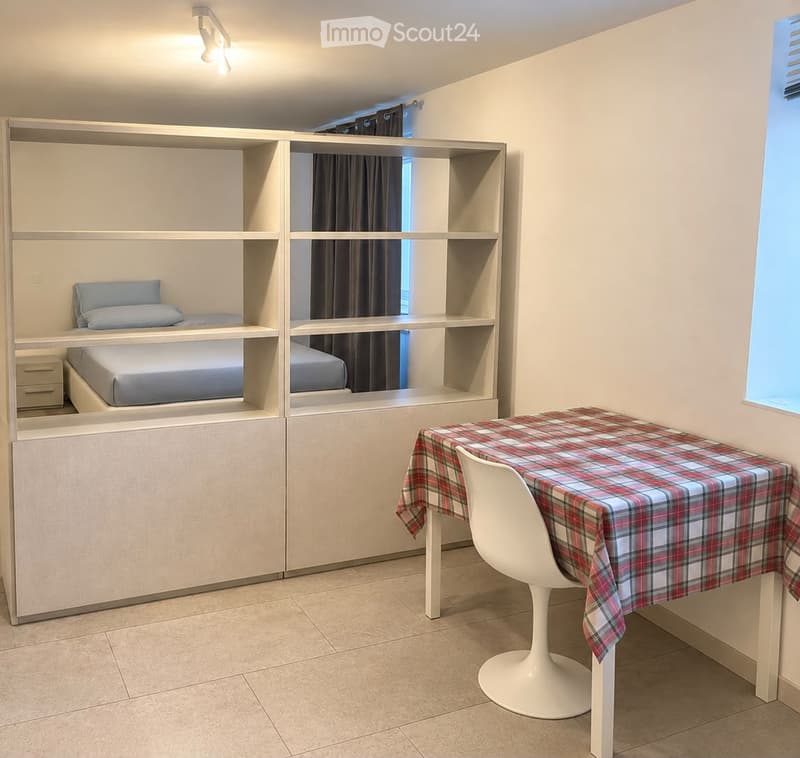 A compact, modern bedroom with built-in bunk beds, a small table with a plaid tablecloth, and a white chair. The room features light-colored walls, tiled flooring, and built-in shelving units.