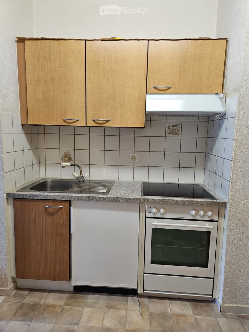 Compact kitchen with wooden cabinets, a sink with a faucet, an electric stove, and an oven. The walls are tiled, and the floor is tiled as well.