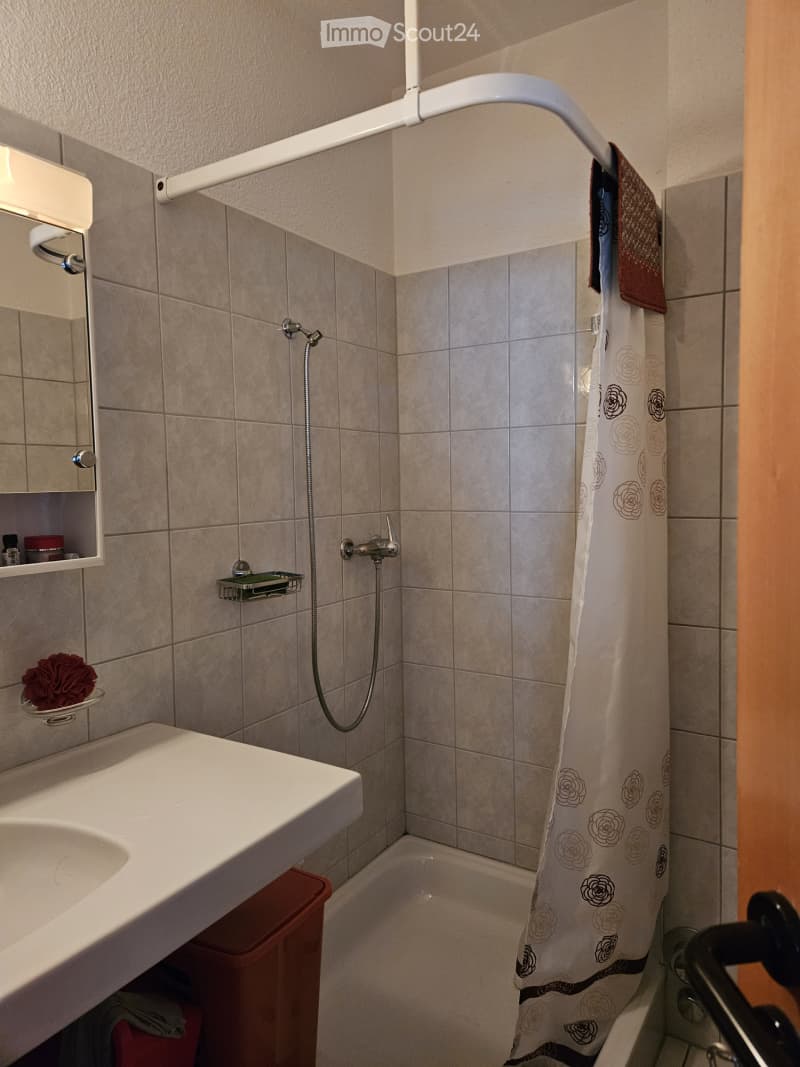Bathroom with a white sink, mirror, bathtub, and shower area with a curtain. Beige tiled walls and floor, towel rack with a towel, and a small shelf with toiletries.
