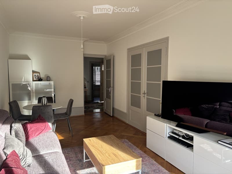 Living room with a sofa, coffee table, television, dining area with chairs and table, and a cabinet with decorative items.