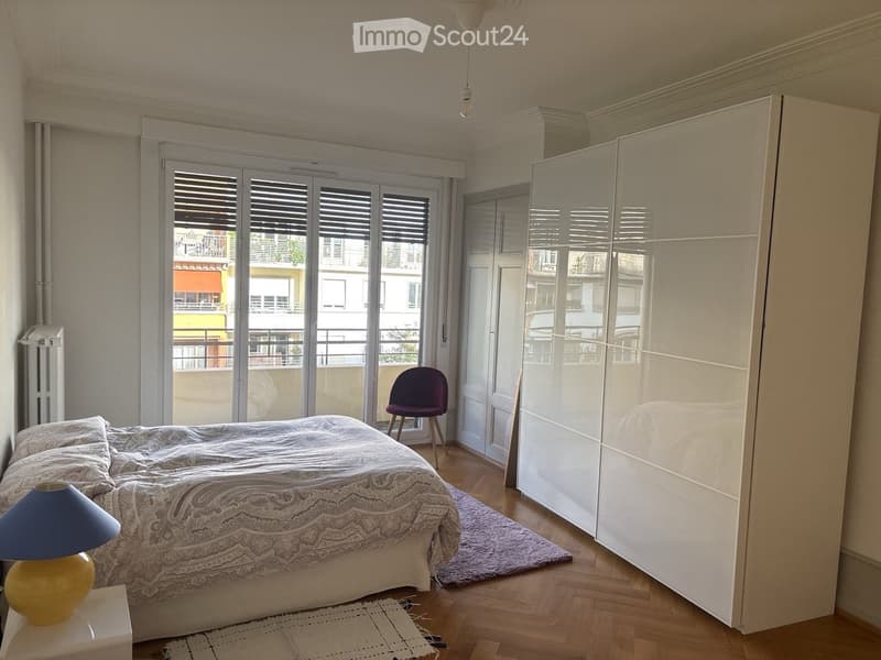 A well-lit bedroom with a large bed, nightstand with a lamp, a purple chair, and a large white wardrobe. The room features wooden flooring and large windows with shutters leading to a balcony.