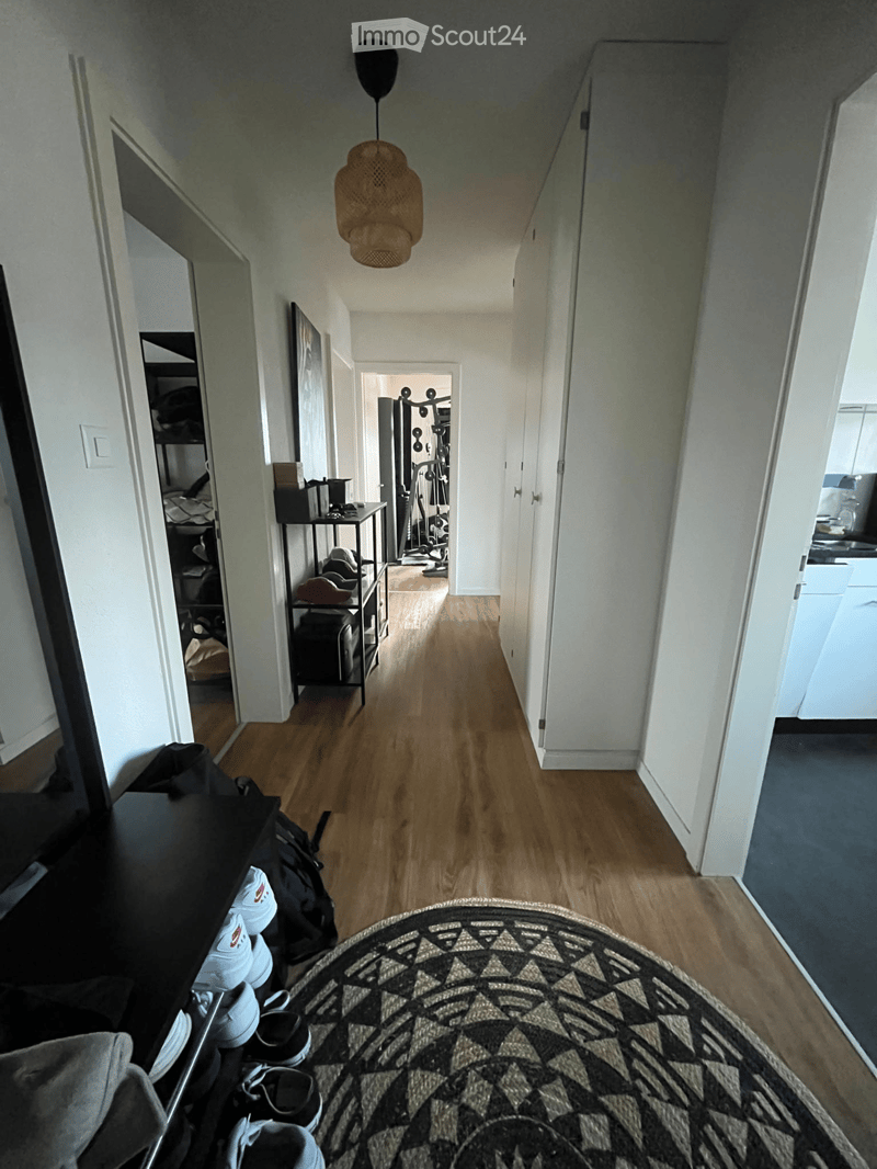 A long, narrow hallway with wooden flooring, white walls, and a hanging woven light fixture. The hallway features several open doors leading to different rooms, including a bedroom, a gym area, and a kitchen. A small side table with items and a shoe rack