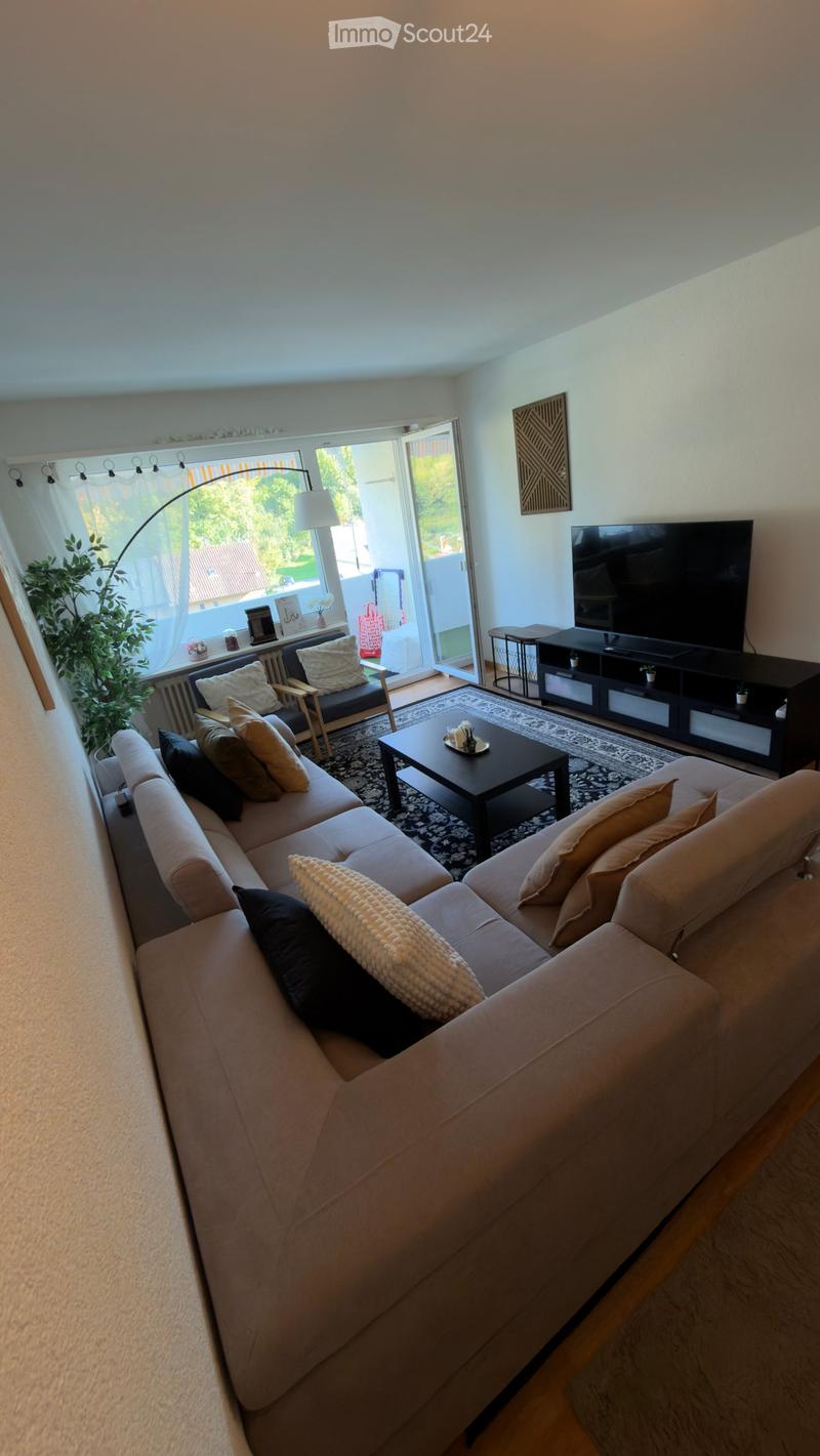 Modern living room with a large sectional sofa, coffee table, armchair, TV stand, floor lamp, and a plant near the window. The room features large windows with sheer curtains and a view of greenery outside.