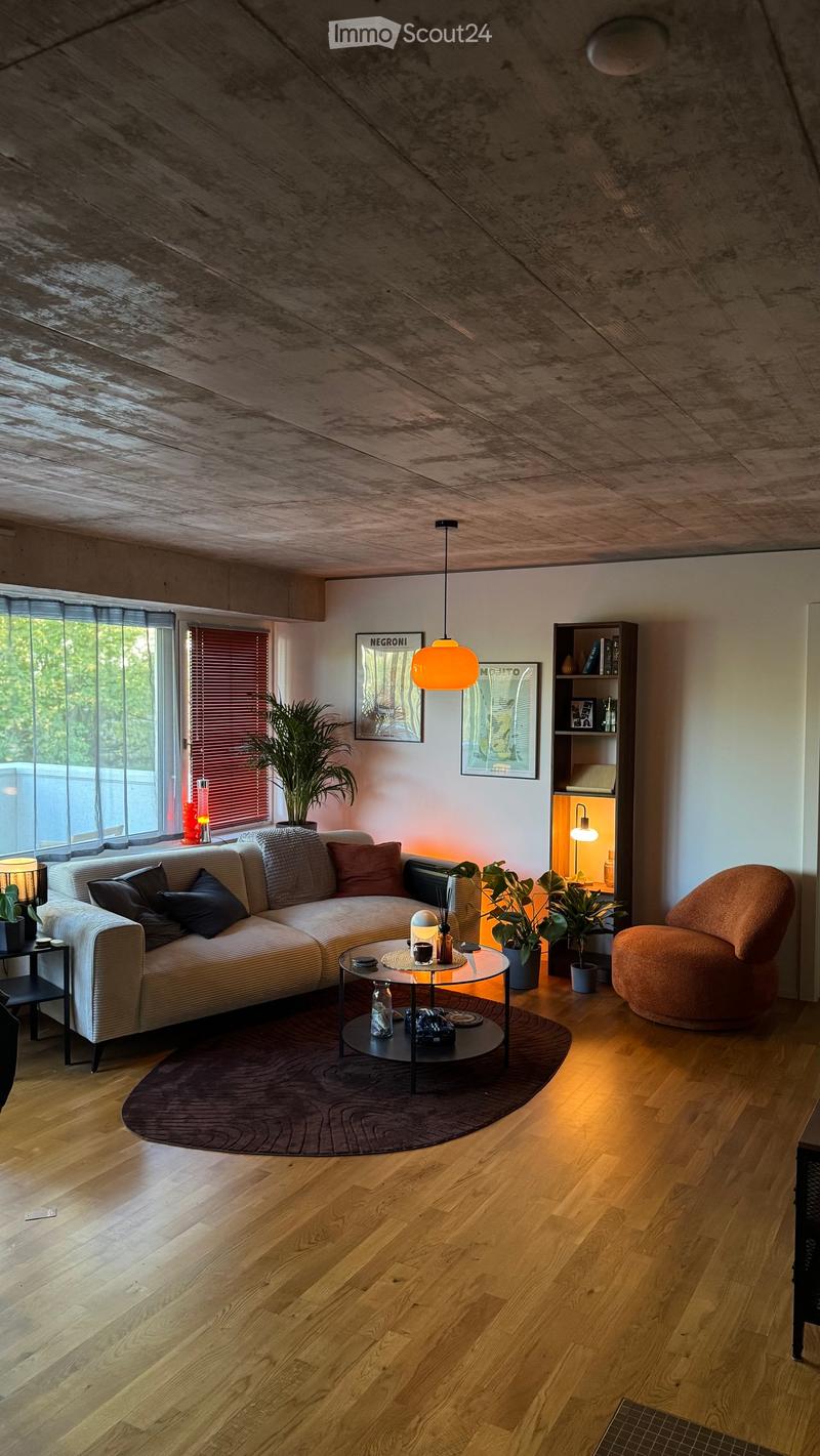 Modern living room with wooden flooring, a sectional sofa, a round glass coffee table, a side table, a large potted plant, a bookshelf, and a hanging lamp.