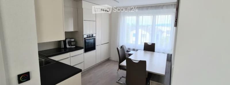 Modern kitchen and dining area with white cabinets, black countertop, built-in oven, coffee maker, and a dining table with four chairs.