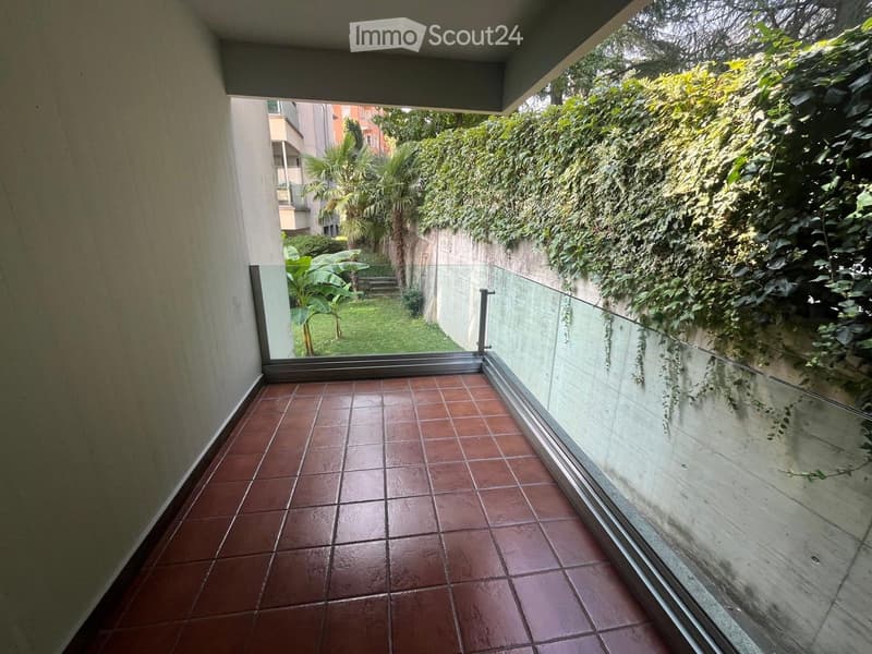 Balcony with terracotta tiles, glass railing, and view of a garden with greenery and trees.