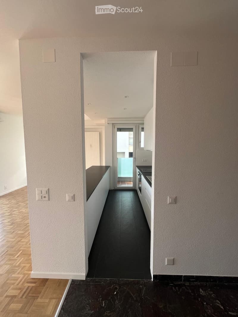 Empty hallway with wooden flooring, white walls, and a view into a modern kitchen area with dark countertops and white cabinets.