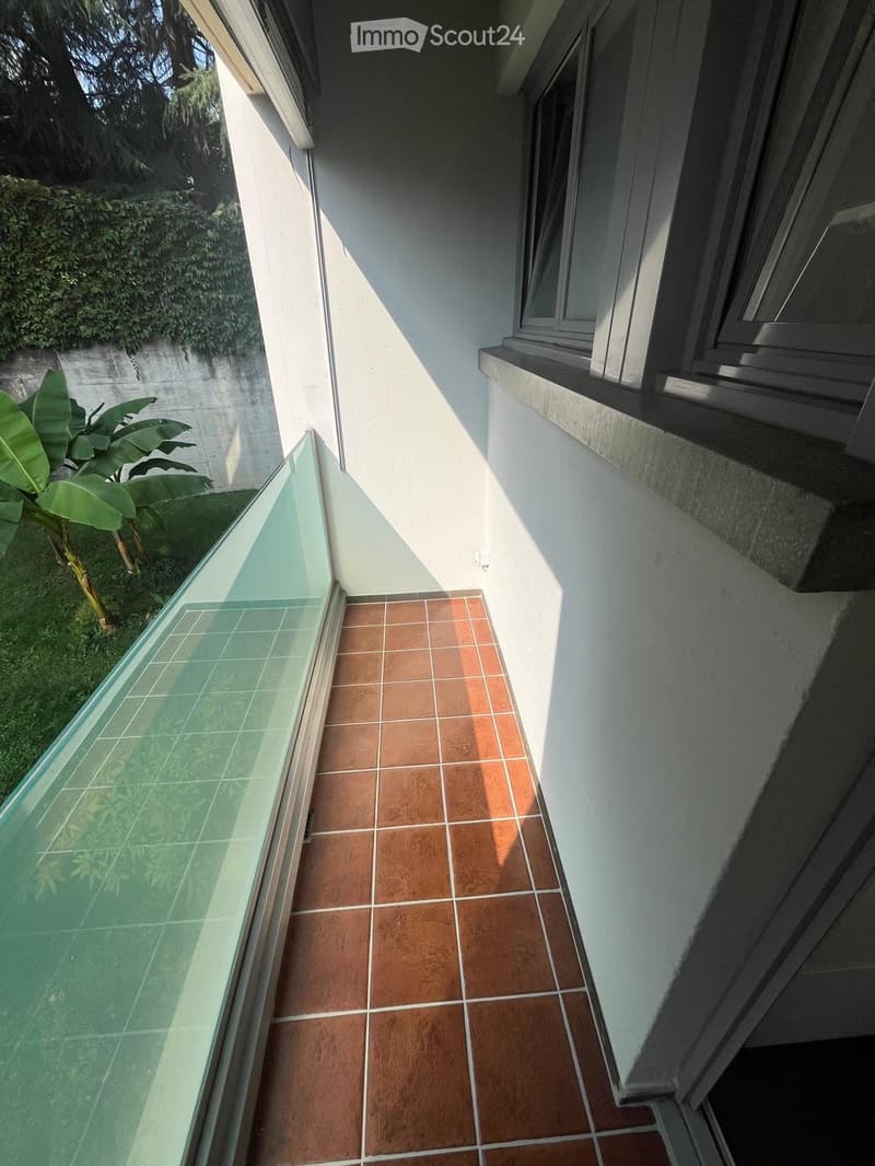 Balcony with glass railing, terracotta tiles, and a view of greenery.