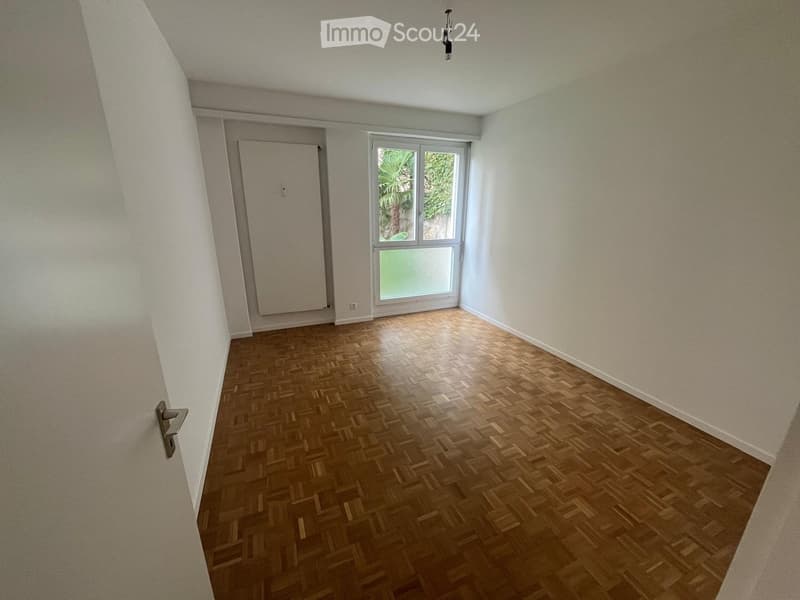 Empty room with wooden parquet flooring, white walls, and a large window with a view of greenery outside.